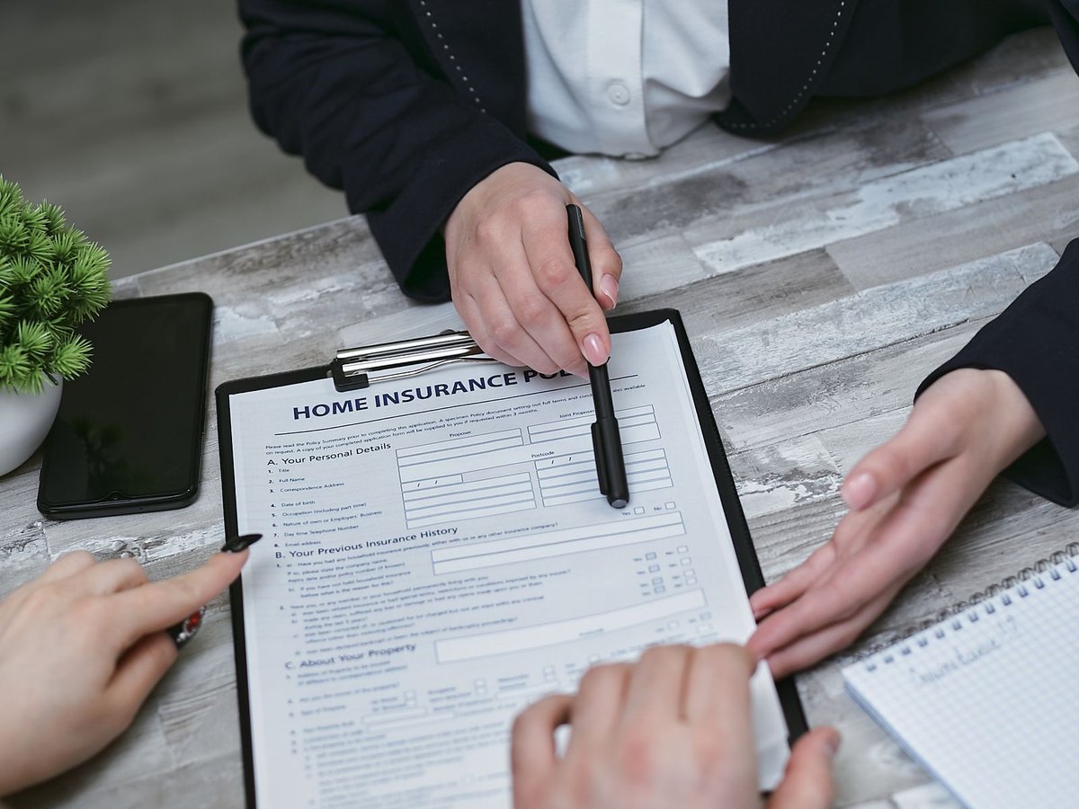 An insurance agent points to a home insurance policy application on a clipboard while two homeowners review the coverage details