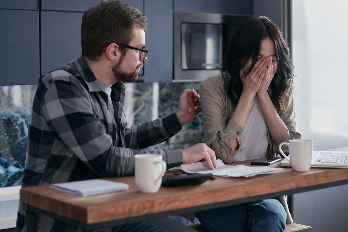 Couple at a kitchen table reviewing bills and financial paperwork with a laptop and coffee mugs, one of them looking stressed