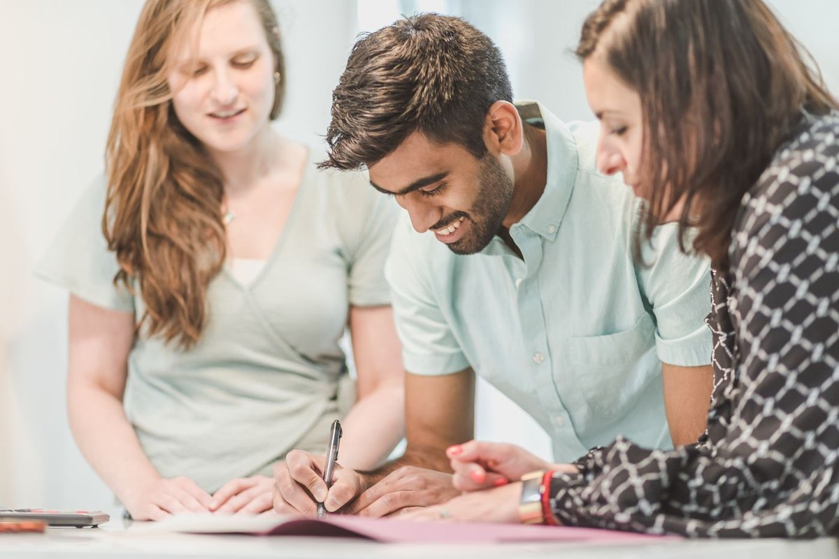 Young man signing mortgage or purchase documents at a table while two colleagues review the paperwork