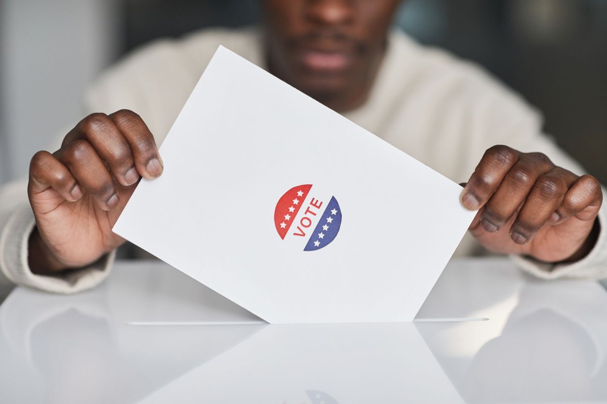 Voter placing a paper ballot marked with a vote sticker into a white ballot box
