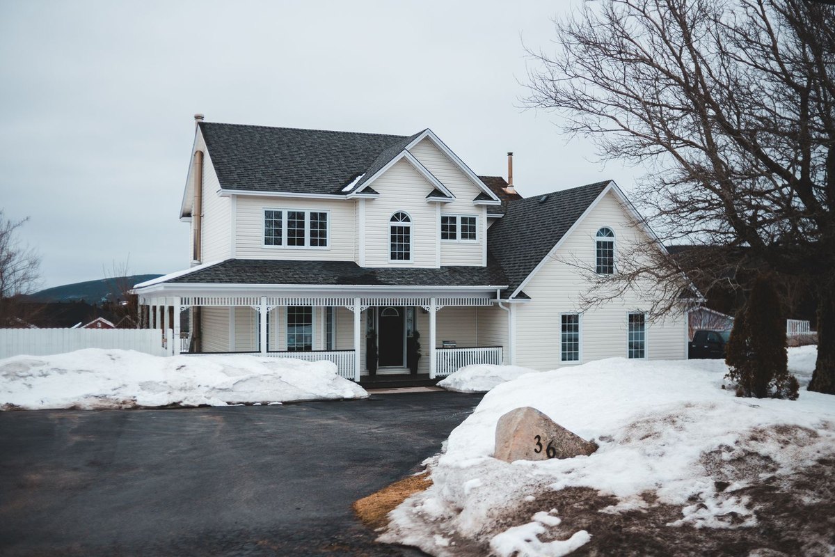 White two-story house with a covered porch and dark roof beside snow-covered lawn and bare trees