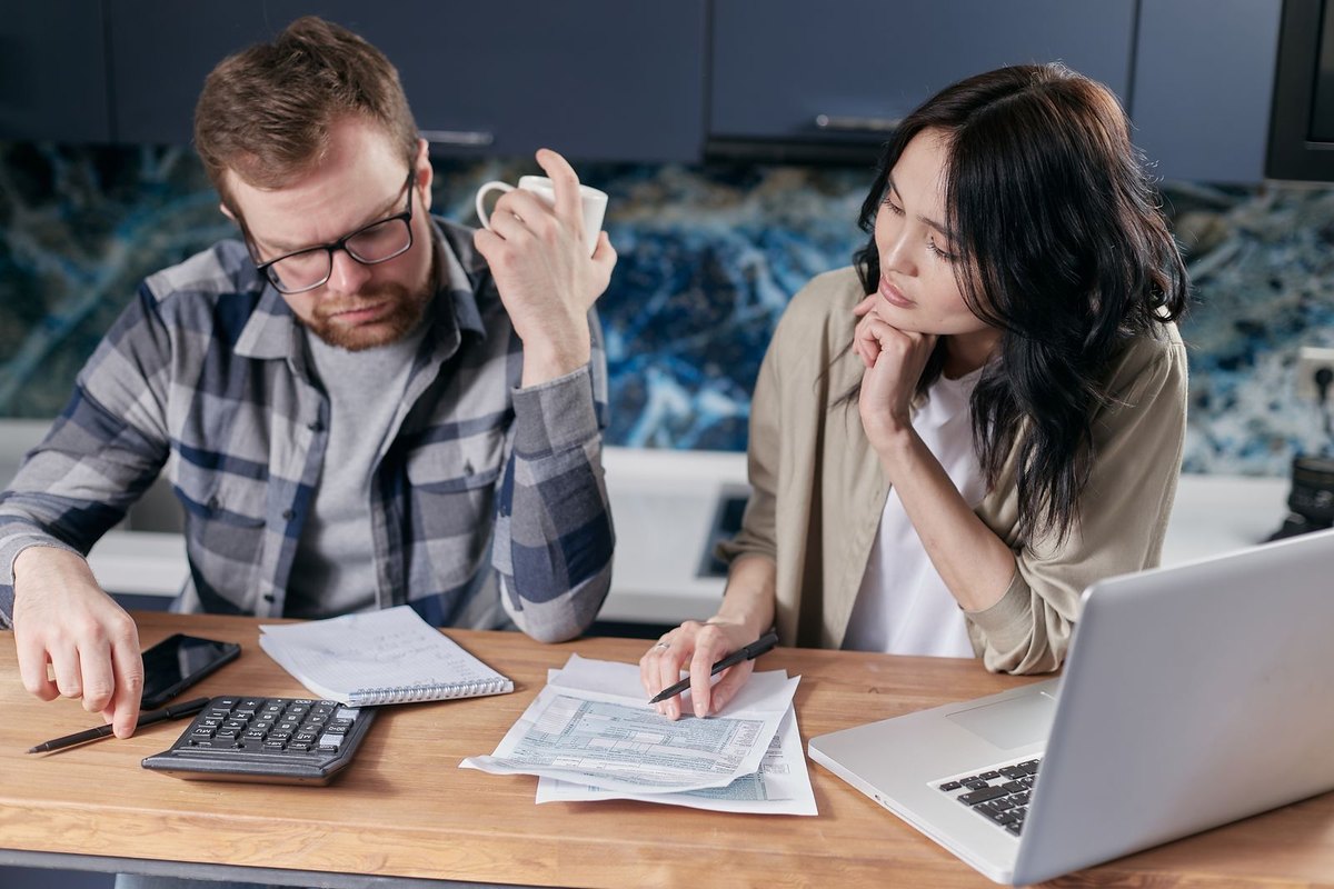 Couple at a kitchen table reviewing paperwork with a calculator and laptop, the man resting his head on his hand while the woman looks at him