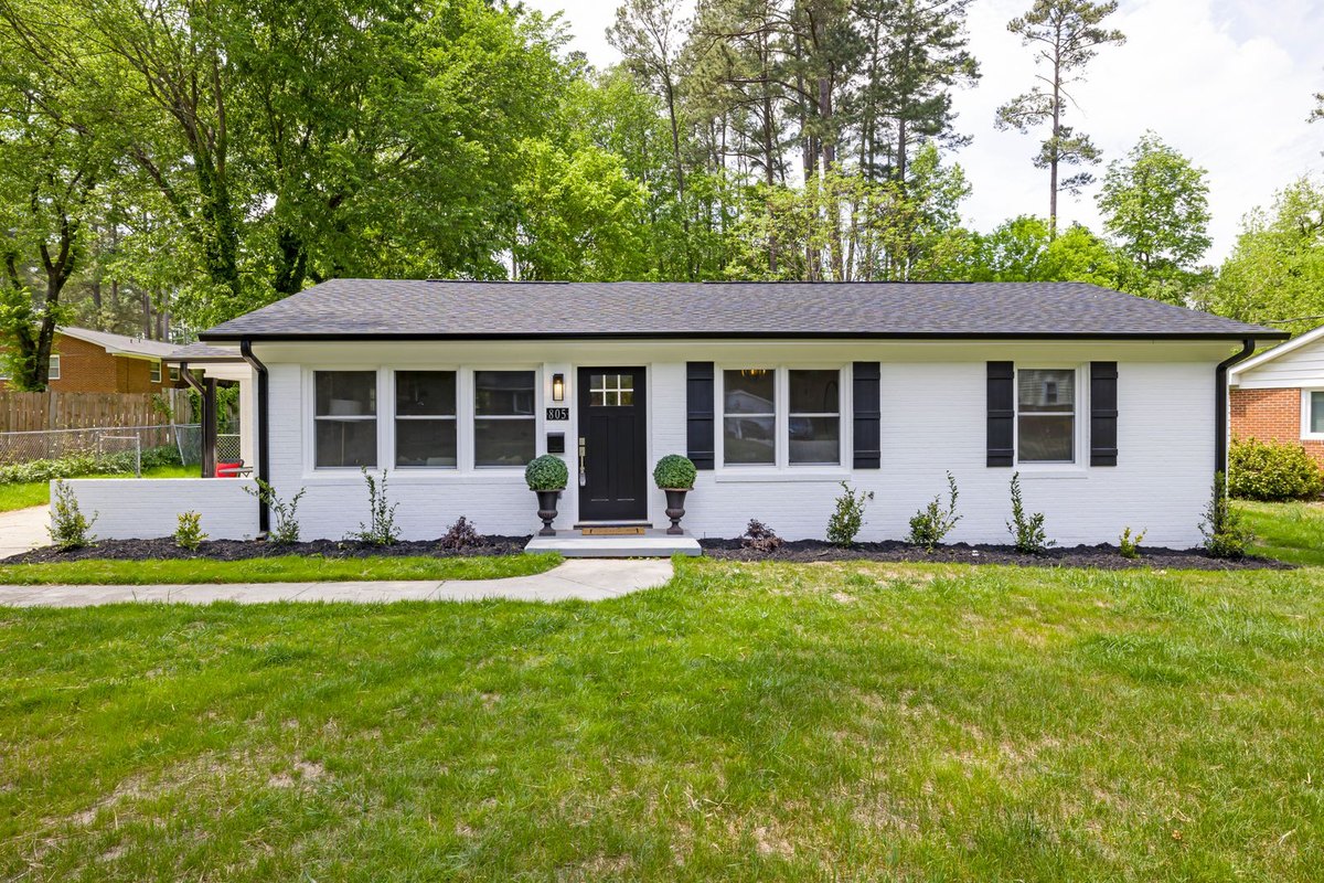 Single-story white ranch house with black shutters, a wide green lawn, and tall trees in a quiet suburban neighborhood
