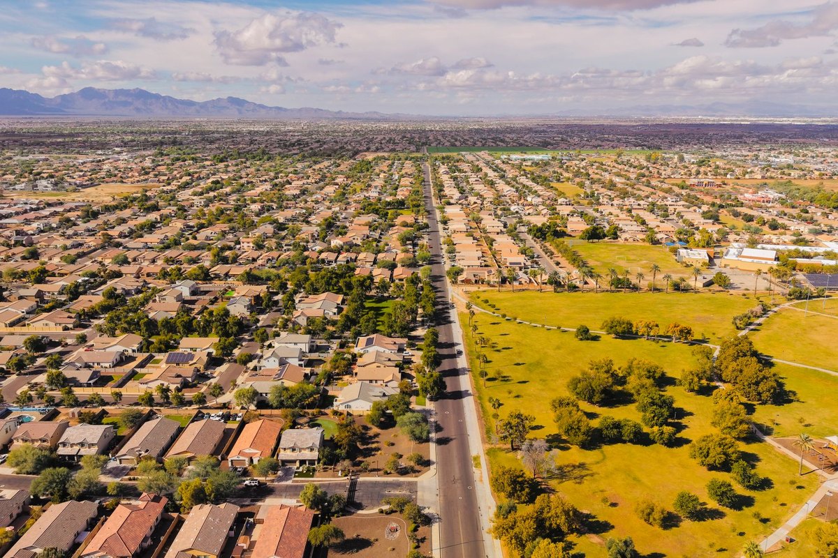 Aerial view of a Phoenix-area suburban neighborhood with rows of tile-roof homes, a wide arterial road, and open desert lots under a clear sky