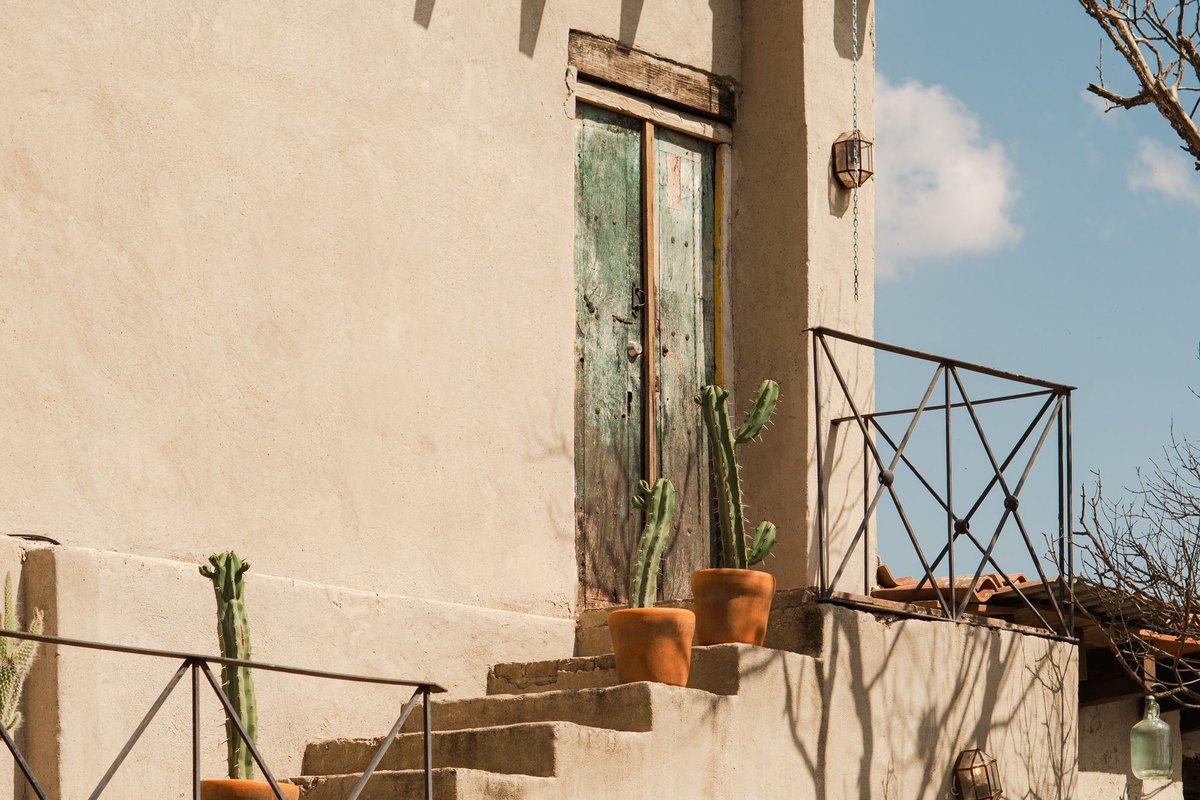 Sunlit adobe-style home with a flat roof, wooden beams, an exterior staircase, and potted cacti along the walk