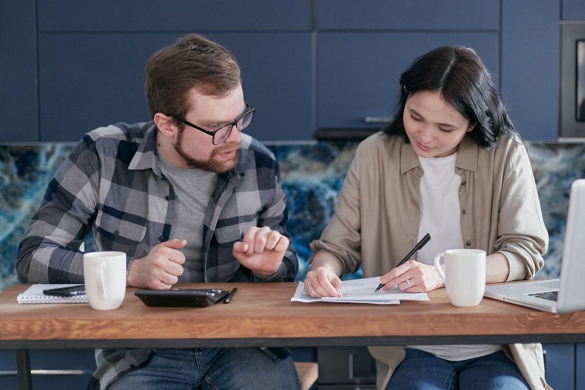 Man and woman sitting at a wooden table with a laptop, calculator, coffee mugs, and paperwork, comparing loan options