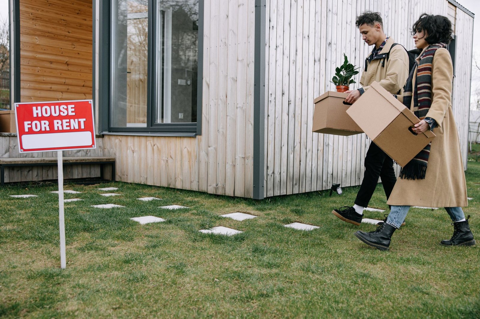 Couple carrying moving boxes past a sign in front of a modern home