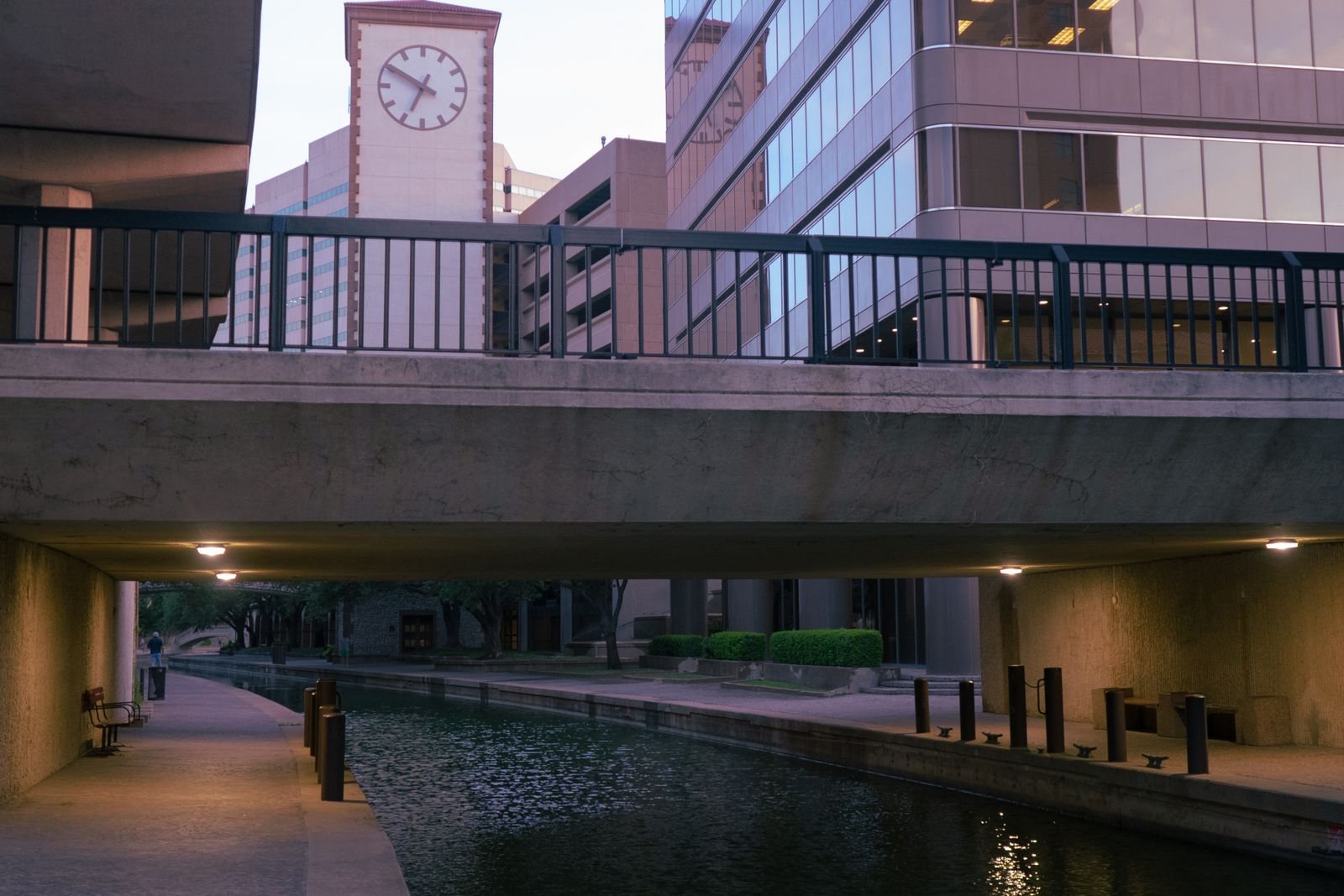 Canal walkway under a bridge beside office buildings and a clock tower