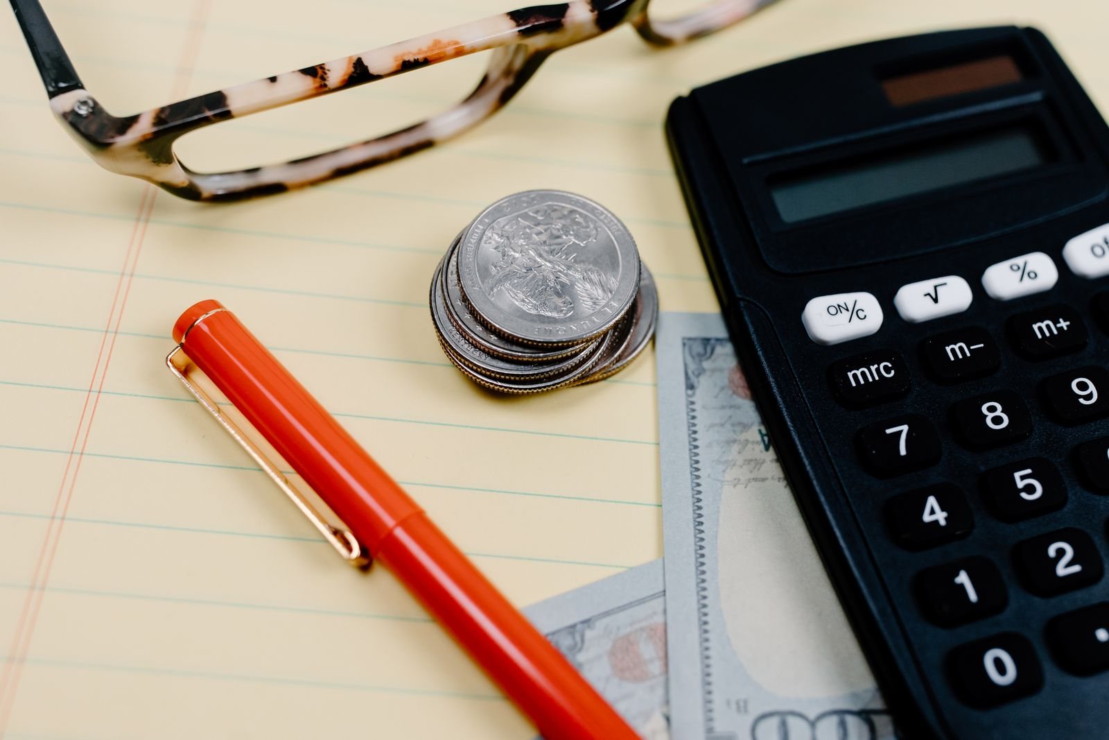 Calculator, stacked coins, eyeglasses, and a red pen on a notepad