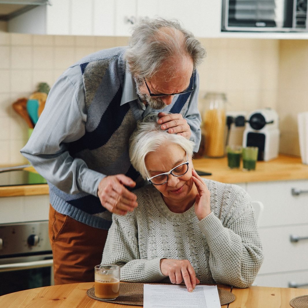 Older couple in their kitchen, the husband leaning over his wife as she reads a document at the table