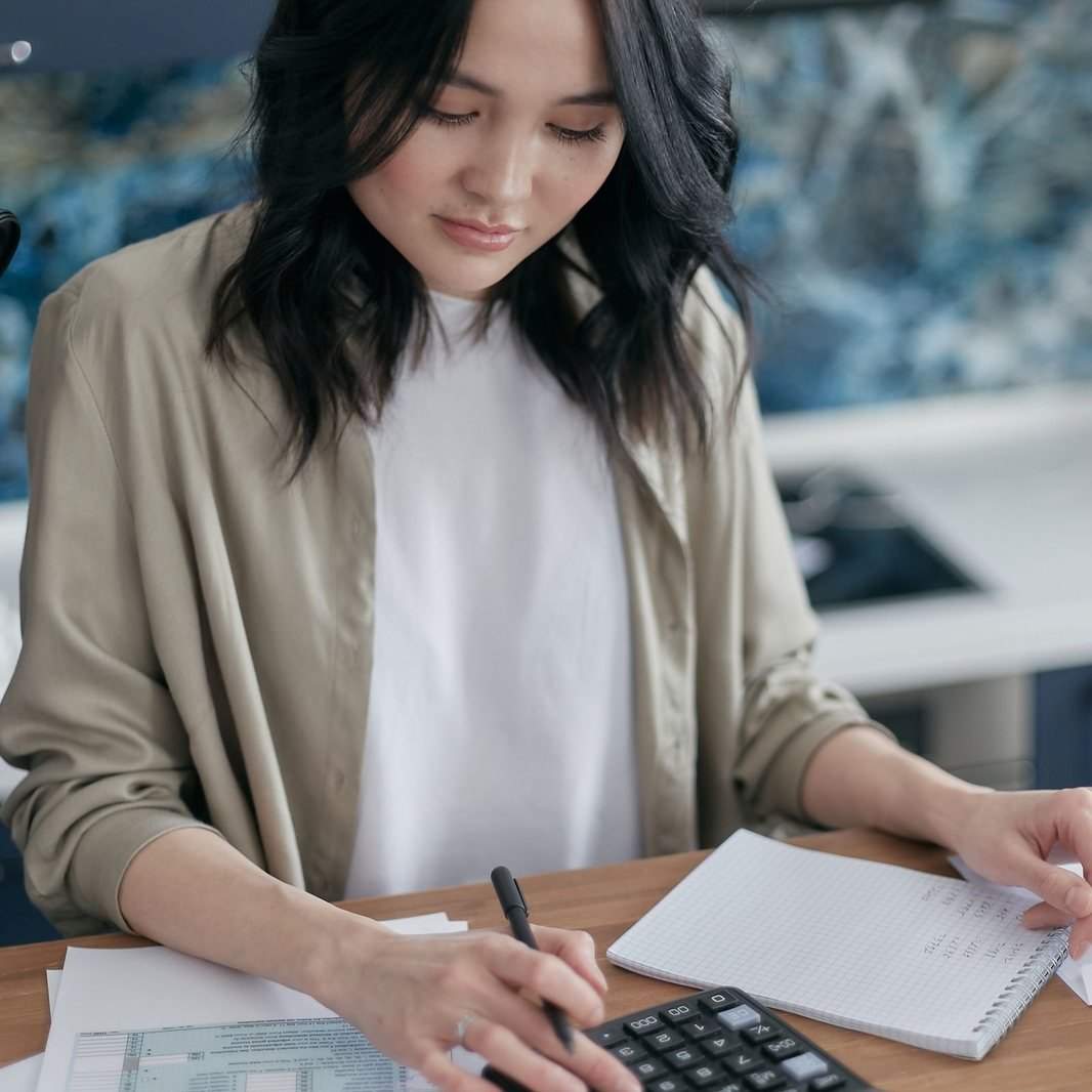 Woman at a kitchen table using a calculator and laptop while reviewing household paperwork